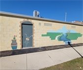 Partially completed mural painted on the side of a tan brick building. On the left, a blue and red patterned vase with flowers is painted next to a doorway where a bicycle is outlined. On the right, a large mural depicts a river curving through green fields under a blue sky, with a tree on the horizon. The artwork is still in progress.